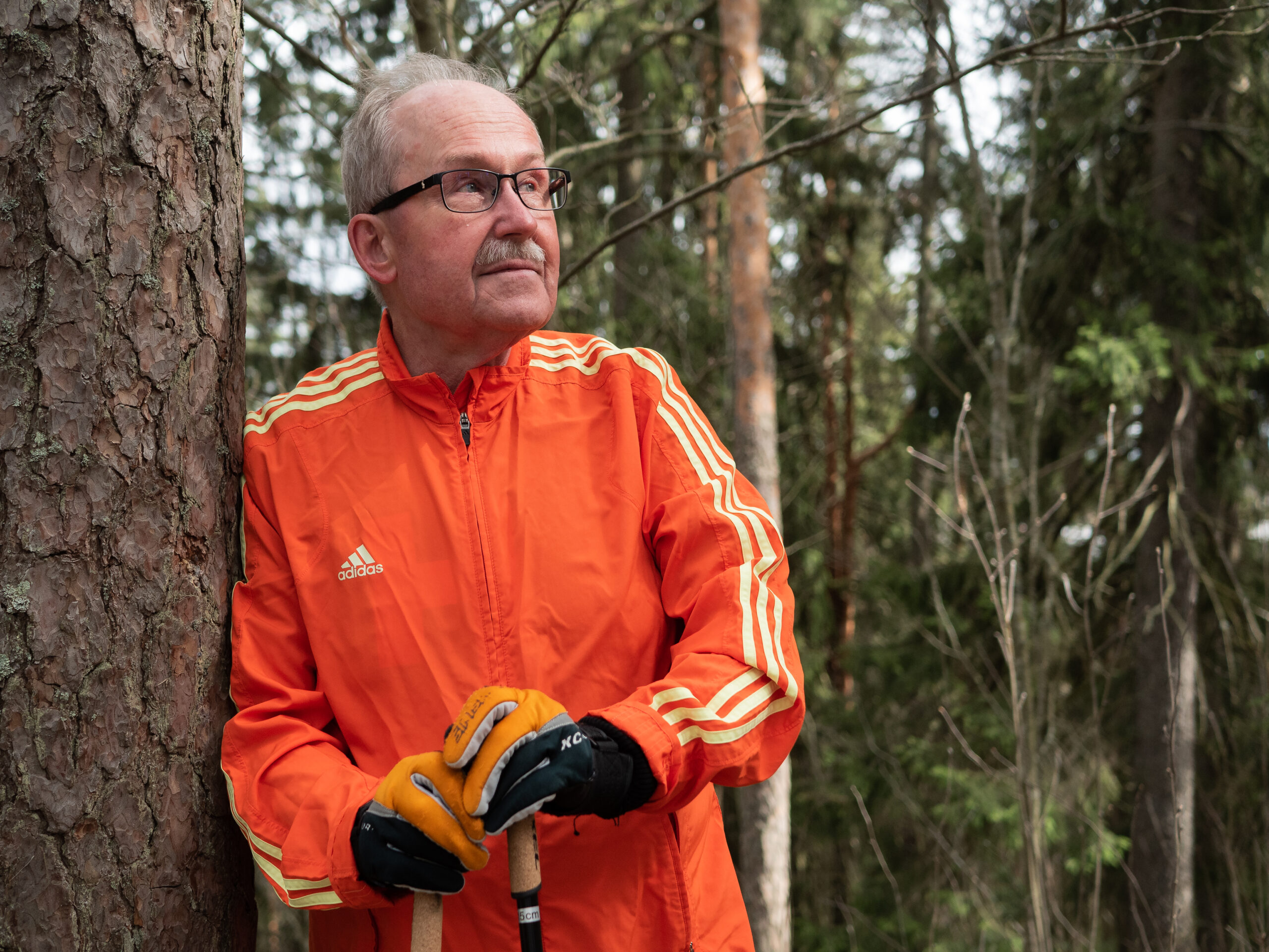 Lahden Talojen Kotoilija Risto Adidaksen verkkatakissa sauvakävelyllä, nojaa puuhun ja katsoo yläoikealle tyytyväisenä.