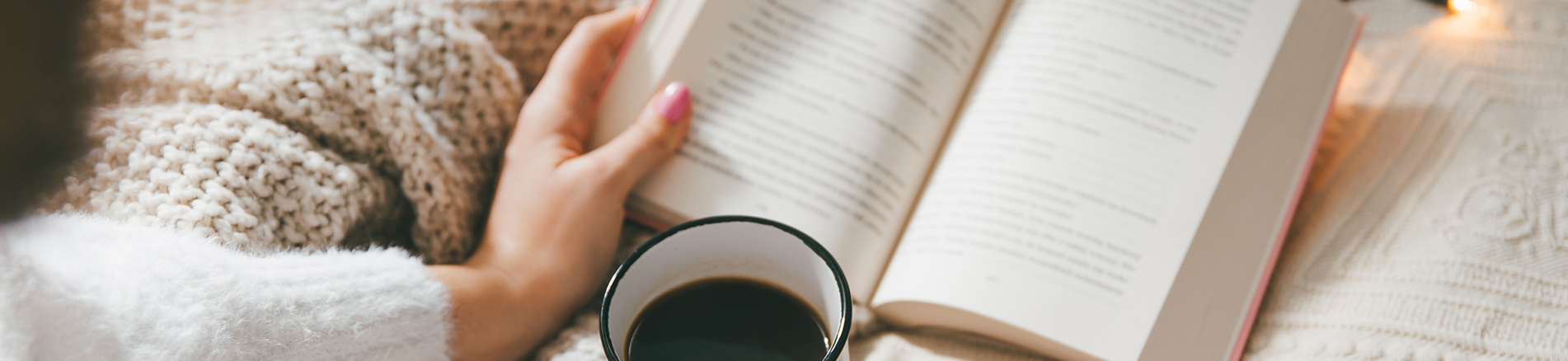 Kuvassa on nainen lukemassa kirjaa ja juomassa mustaa kahvia. Hän on vaalean peiton alla
